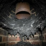 Close-up of heavy, dark, flaky creosote buildup coating the inside walls of a neglected residential fireplace flue, illuminated by a flashlight.